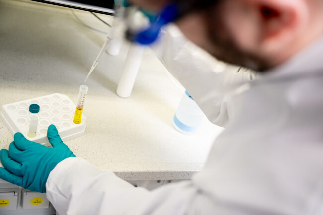 Seorang ilmuwan menambahkan amonia ke dalam botol yang berisi larutan kuning. Botol itu terletak di bangku laboratorium putih. Peneliti mengenakan jas lab putih dan sarung tangan hijau.