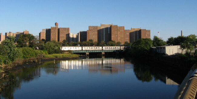 A subway train crossing Coney Island Creek on a sunny afternoon, in front of a cluster of buildings
