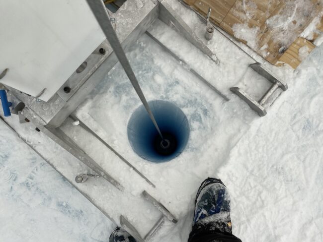 Photo of a long, skinny ice core drill descending into a deep blue borehole.