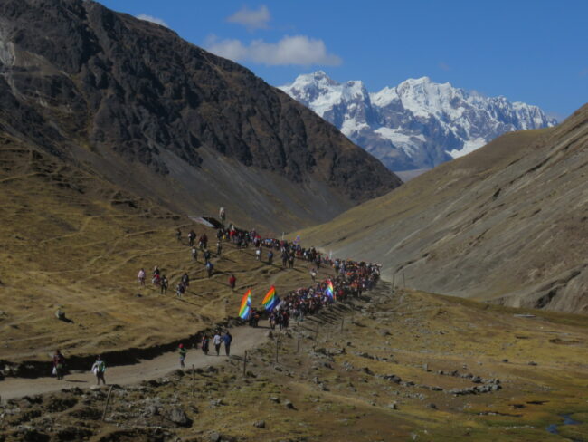 A large group of hikers carrying rainbow flags walks along a mountain trail with snowcapped peaks in the background
