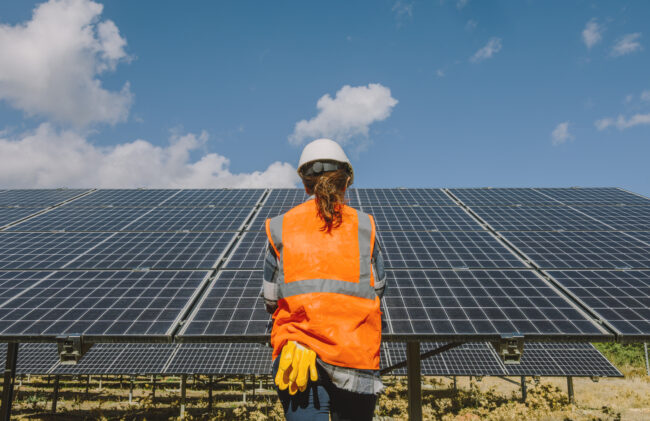 Back view of an engineer standing and working beside solar panels in a sun power station farm. 