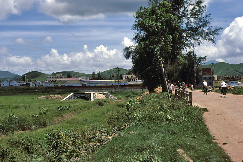 A dirt road with cyclists beside a grassy field, trees, and buildings under a cloudy sky. Shenzen in 1980