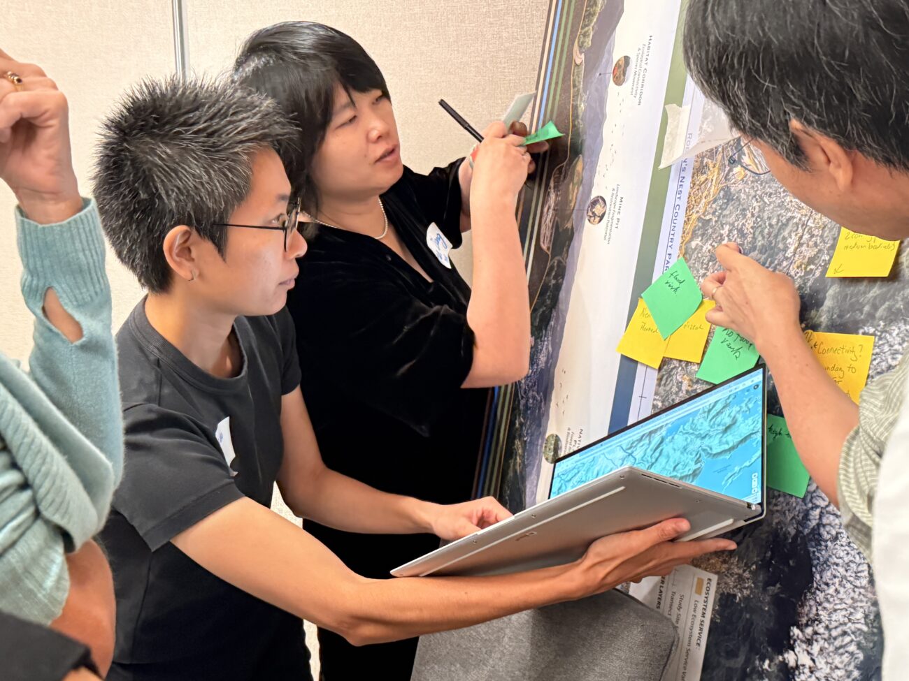 People collaborating around a wall map, adding sticky notes and using a laptop to review geographic data