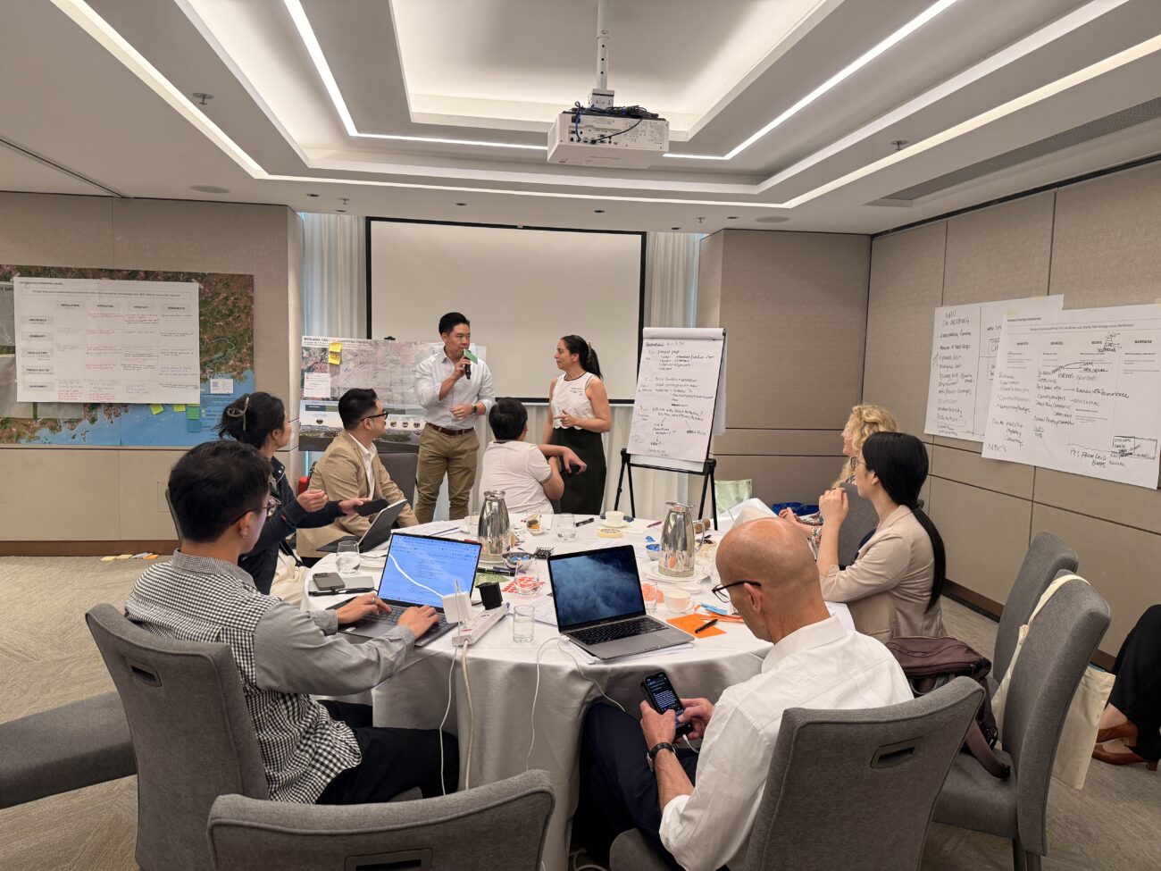 People in a workshop discussing ideas around a table with laptops and flip charts in a conference room