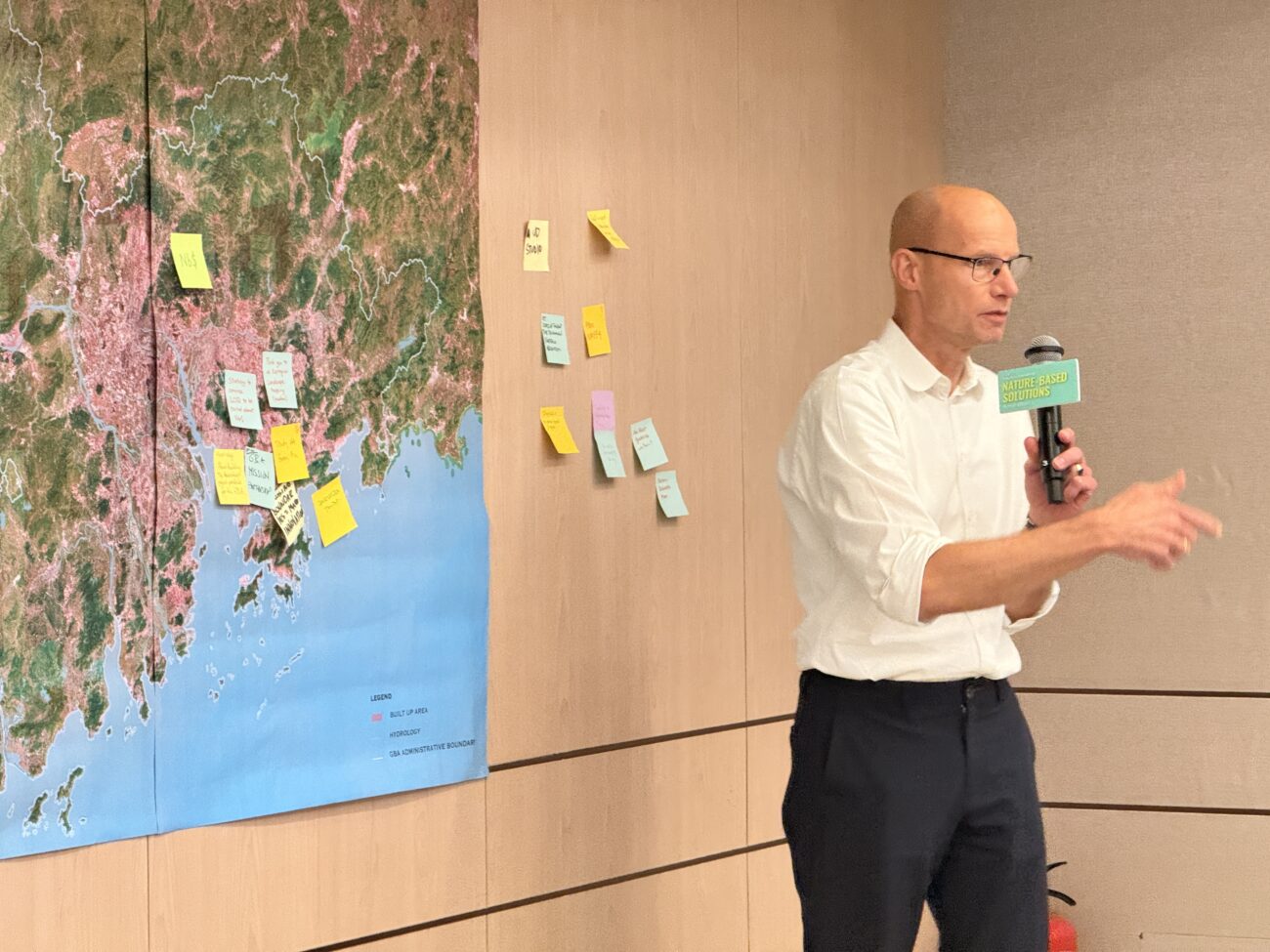 Man speaking into a microphone beside a wall map covered with sticky notes during a presentation