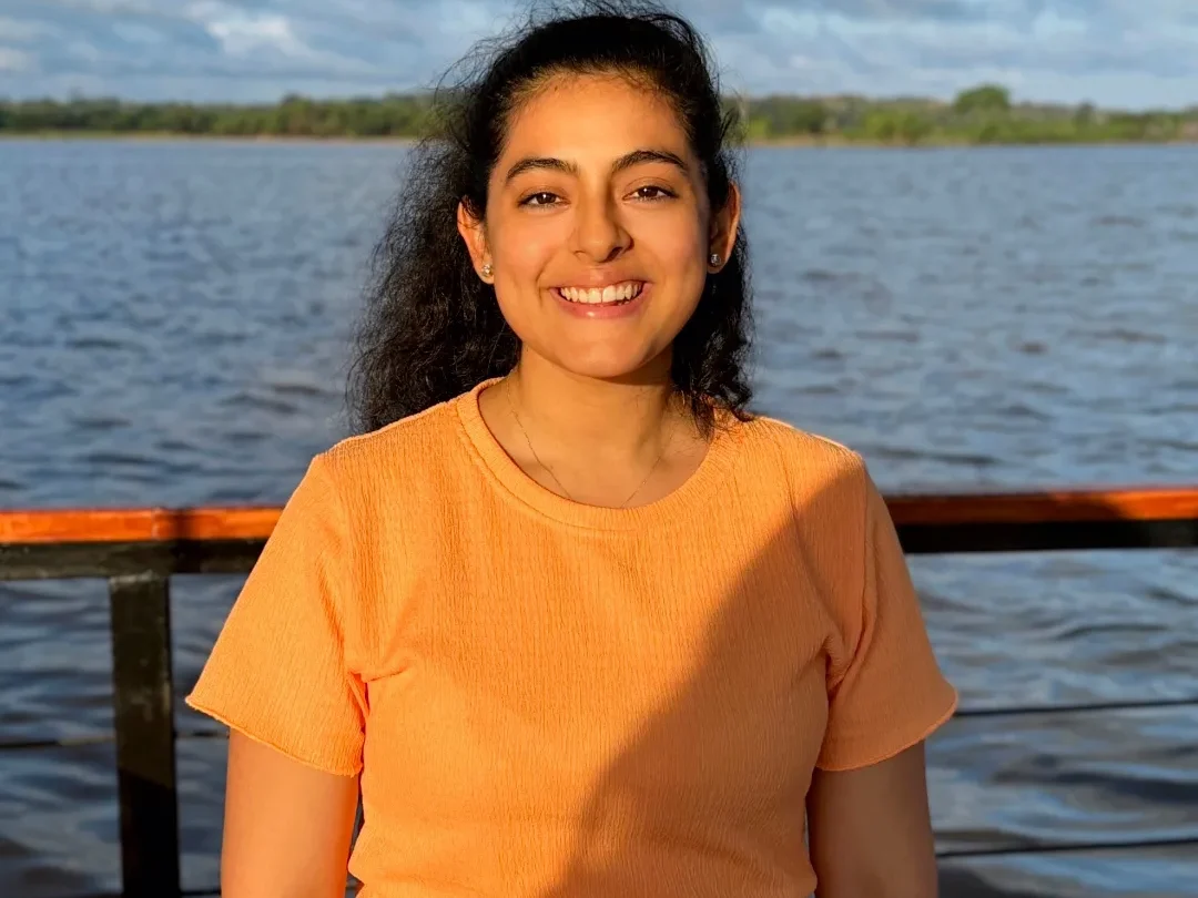 Smiling woman in an orange shirt stands beside a lake under a cloudy sky