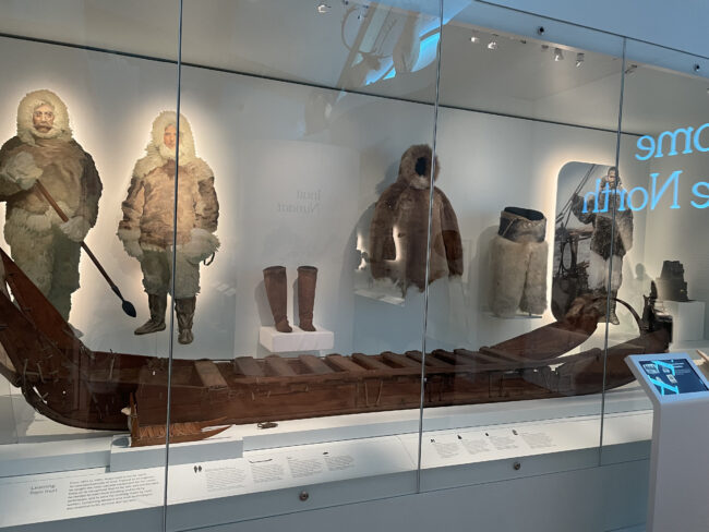 Museum exhibit displaying Inuit parkas, kayaks, and hunting equipment in a glass case