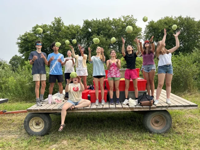 A group of students tosses cabbages in the air while posing on a wagon outdoors