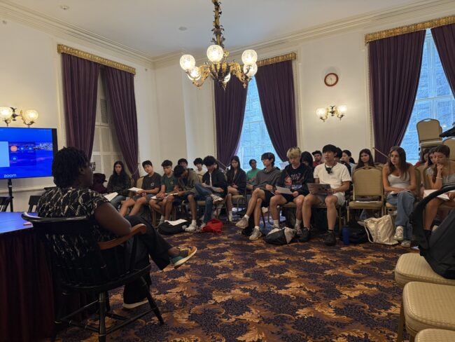 Students sit in an ornate room facing a presenter and a Zoom screen