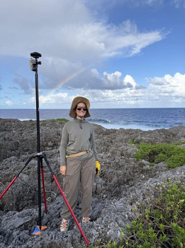 A woman stands beside surveying equipment on a rocky coastline, with ocean waves and a rainbow behind her