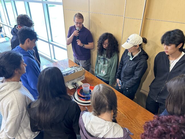 A student group gathers around a table as a man speaks into a microphone