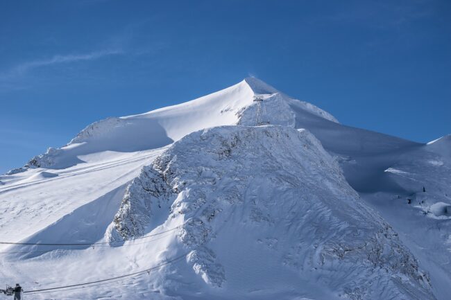 Snow-covered mountain peak with cable car lines and a tower beneath a clear blue sky