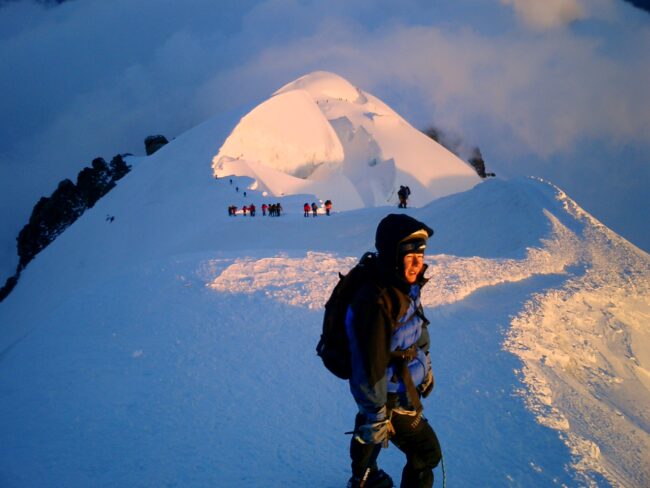 Mountaineer in winter gear climbing a snow-covered peak, with a group of climbers behind him