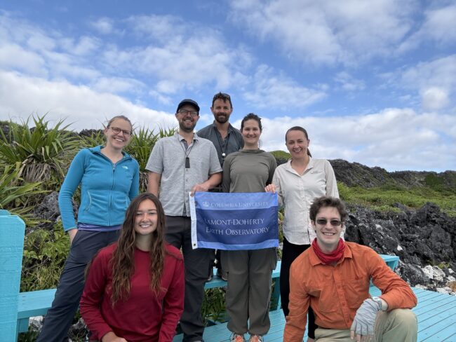 Seven people pose outdoors with a Columbia University Lamont-Doherty Earth Observatory flag