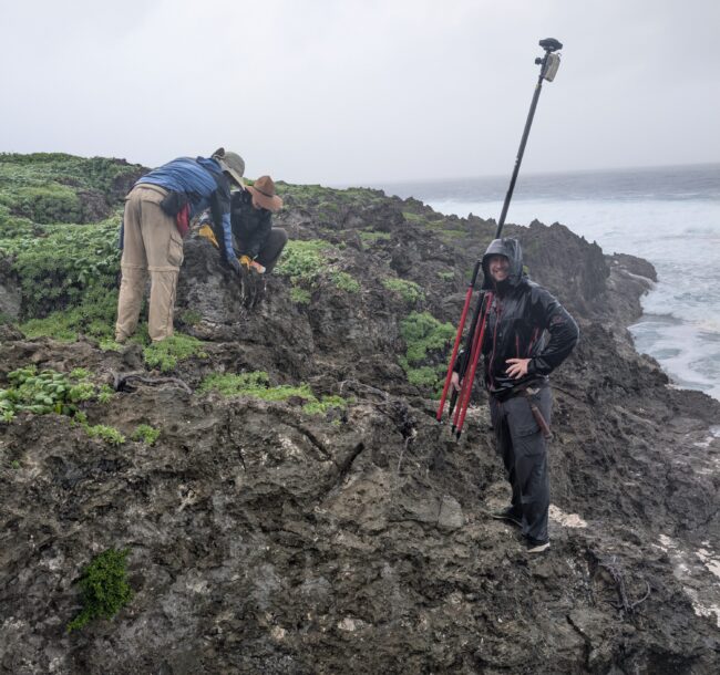 Three people in rain gear work on a rocky coastline beside the ocean, using surveying equipment