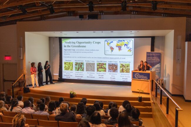 Three presenters discuss opportunity crops in a greenhouse as an audience views a slide on climate-resilient crops