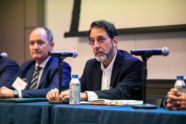 Two men sit at a panel table with microphones, water bottles, and notebooks