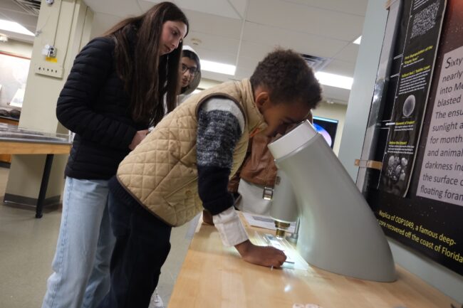 Child looks into magnifier at sediment cores with adults in background