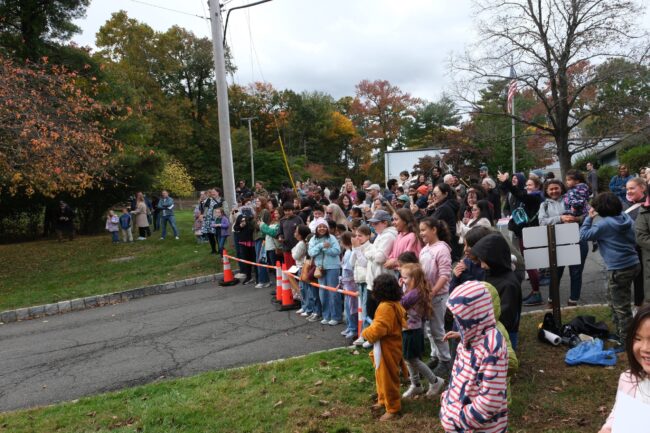 A large crowd of adults and children gathers outdoors in autumn, watching an event along a residential street