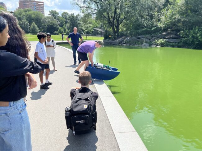 People watch as a man launches a small blue remote-controlled boat into a green pond in a city park