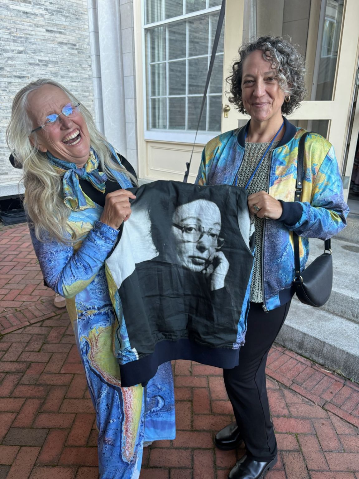 Robin Bell and Vicki Ferrini hold a black-and-white portrait of a bespectacled Marie Tharp outside a building