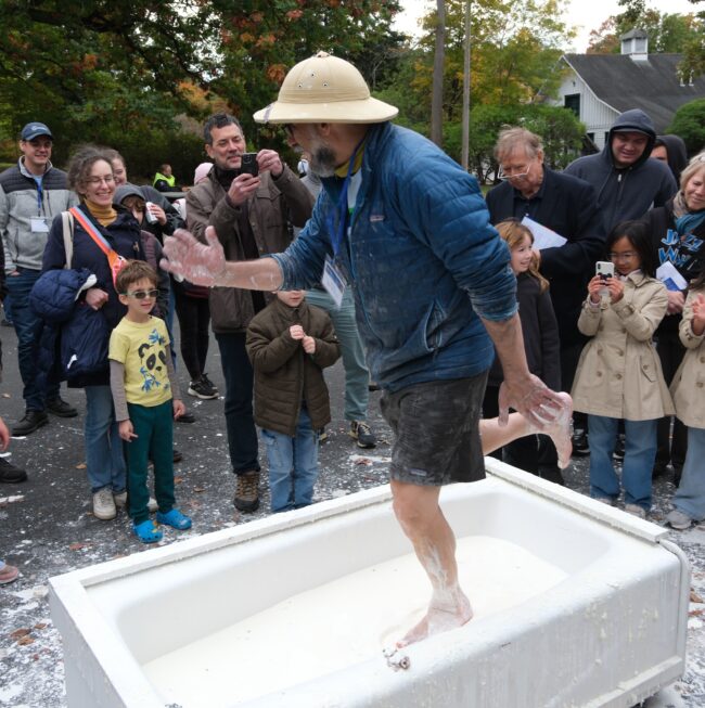 A man in a safari hat steps into a large white tub as a crowd watches and takes photos